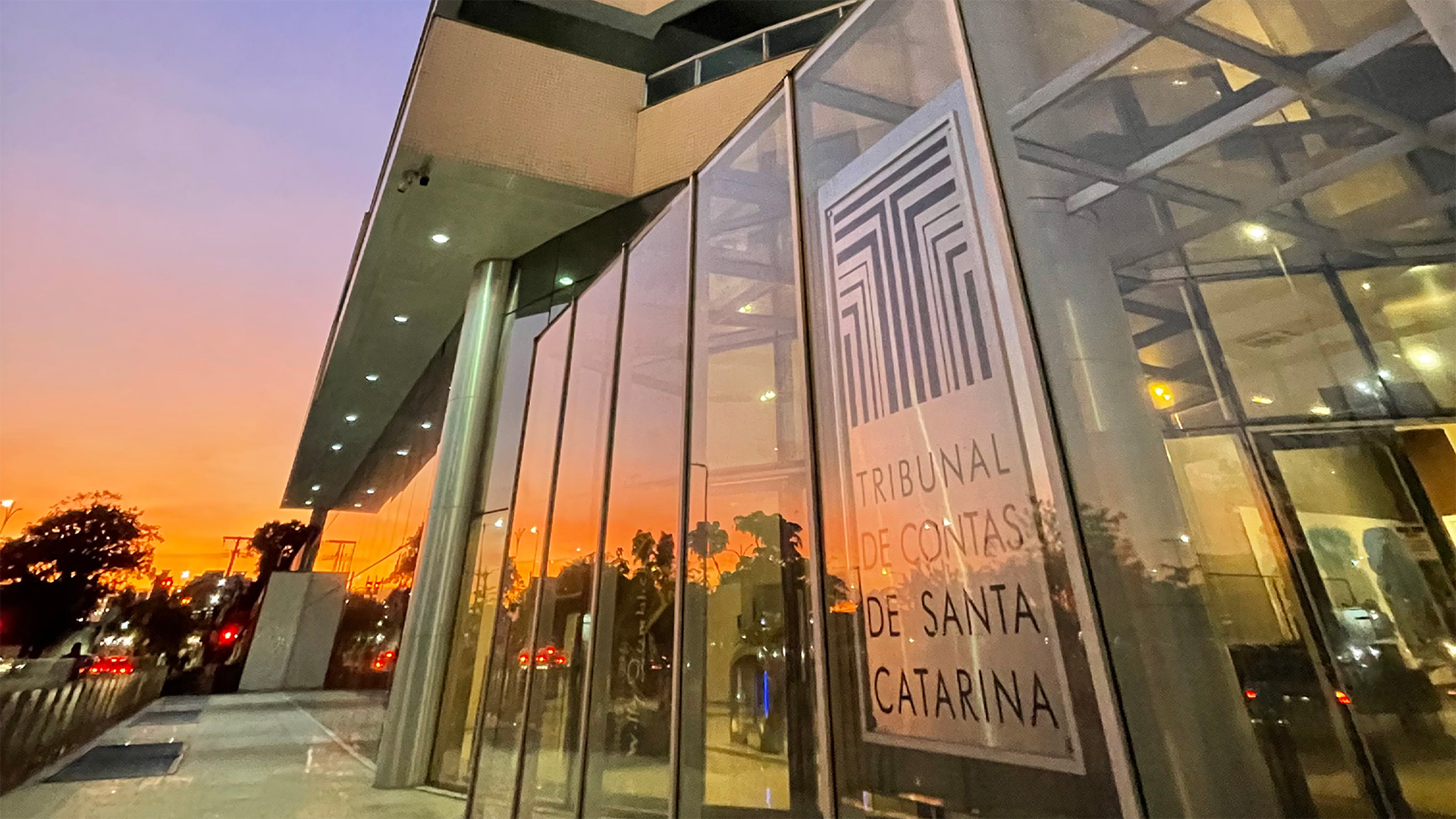 Imagem de parte da sede do TCE/SC, onde aparece parte da logomarca da Instituição, na versão vertical formada pelo desenho de linhas de diferentes espessuras e comprimentos, nas cores bordô e cinza, para construir a letra "T". Abaixo, a inscrição "Tribunal de Contas de Santa Catarina", com fontes na cor preta.